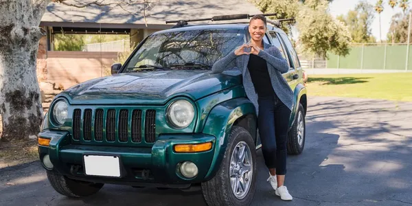 Smiling woman making a heart gesture beside a green SUV outdoors.
