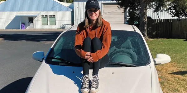 A young woman in casual clothes sits on the hood of a white car, smiling.